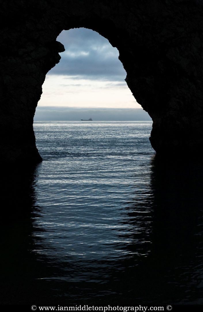 A view through the Durdle Door arch as a cargo ship sails by in the distance, Dorset, England. Durdle door is one of the many stunning locations to visit on the Jurassic coast in southern England.