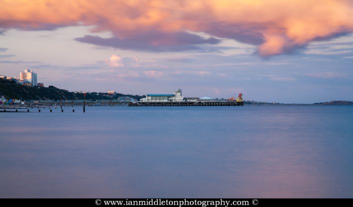 Bournemouth Pier and seafront as a storm cloud drifts over as is illuminated pink by the sunset. Highcliffe can also be seen on the far right.