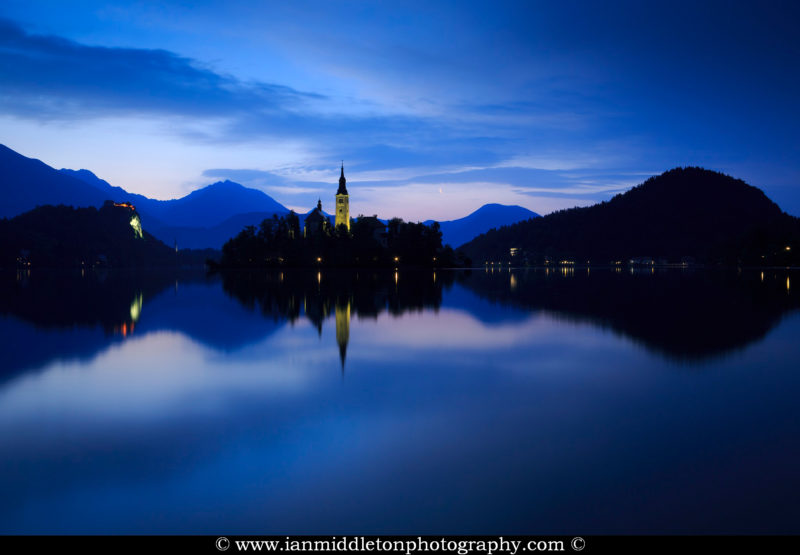 View across the beautiful Lake Bled, island church of the assumption of Mary, and hilltop castle, all topped off with the crescent of the waning moon at dawn, Slovenia.
