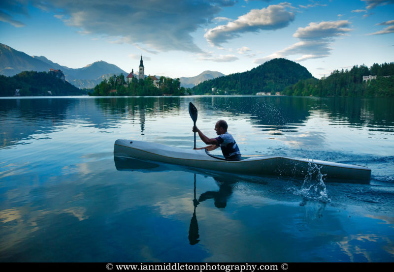 Man canoeing on Lake Bled, Slovenia with the island church of the assumption of saint mary, the clifftop castle in the background.