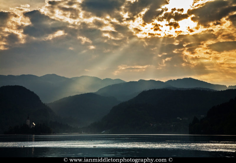 Sun bursting through the clouds and sunrays illuminating the Island Church and mountains in the background, Bled Lake, Slovenia. Some people refer to this beautiful spectacle of nature as angel rays from heaven. Lake Bled is Slovenia's most popular tourist destination.