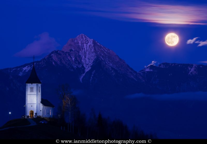 Full moon rising at dusk over Jamnik church of Saints Primus and Felician, perched on a hill on the Jelovica Plateau with the kamnik alps and Storzic mountain in the background, Slovenia.