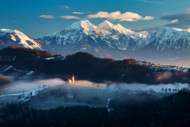 View from Rantovše hill across to Sveti Tomaz nad Praprotnim (church of Saint Thomas) with the Kamnik Alps behind in the Skofja Loka hills, Slovenia.