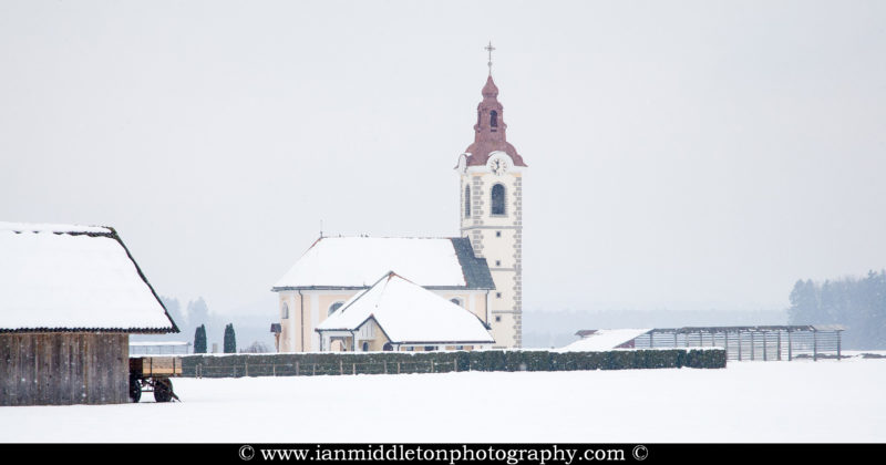 Winter view of the church of saint Simon and Jude as the snow falls, Brnik, near the Ljubljana airport, Slovenia. To the left is a farmer's hut and to the right is a Kozolec, a wooden rack for drying hay in the summer.