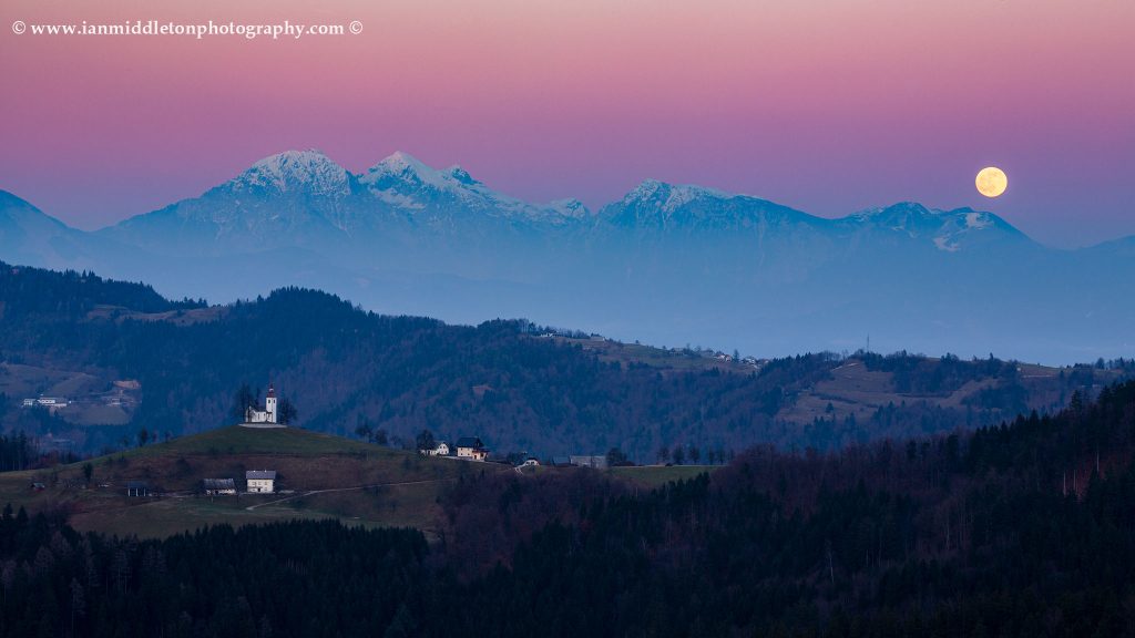 The full moon rising in winter over the mountains behind Sveti Tomaz nad Praprotnim (church of Saint Thomas) and the Kamnik Alps, Slovenia.