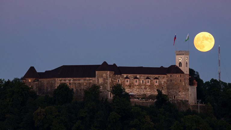 View across the to Ljubljana Castle as the full moon rises beside the tower at sunset. Seen from Tivoli Park, Ljubljana, Slovenia.