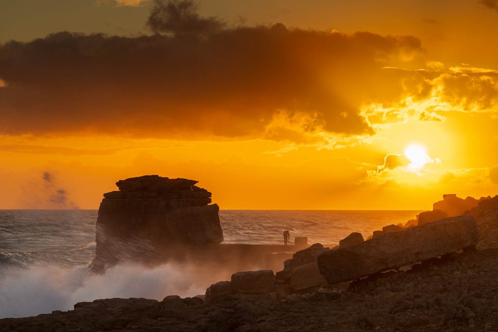 Waves crashing over Pulpit rock at sunset and the promontory at Portland Bill, near Weymouth, Jurassic Coast, Dorset, England. Pulpit Rock is an artificial stack that was left behind after quarrymen dug away a natural arch in 1870s. The Jurassic Coast is a stretch of coastline with a geology that dates back 185 million years, and is a UNESCO World Heritage Site.