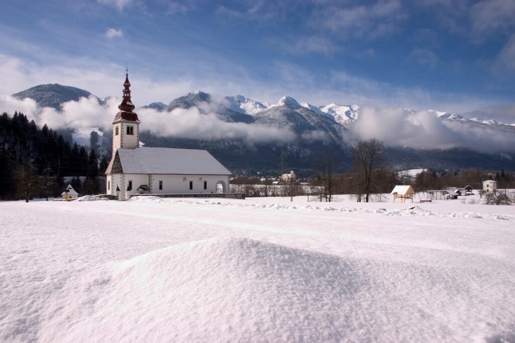 Winter view of the church of the assumption of Mary in Bitnje, a village just outside Bohinjska Bistrica, Bohinj valley, Triglav National Park, Slovenia. You can also see the Church of Saint Nicholos in Bohinjska Bistrica in the background.