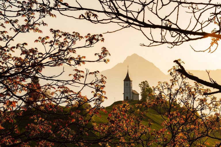 Sunrise at Jamnik church of Saints Primus and Felician, perched on a hill on the Jelovica Plateau with the kamnik alps and Storzic mountain in the background, Slovenia. Framed by some trees in Spring Bloom.