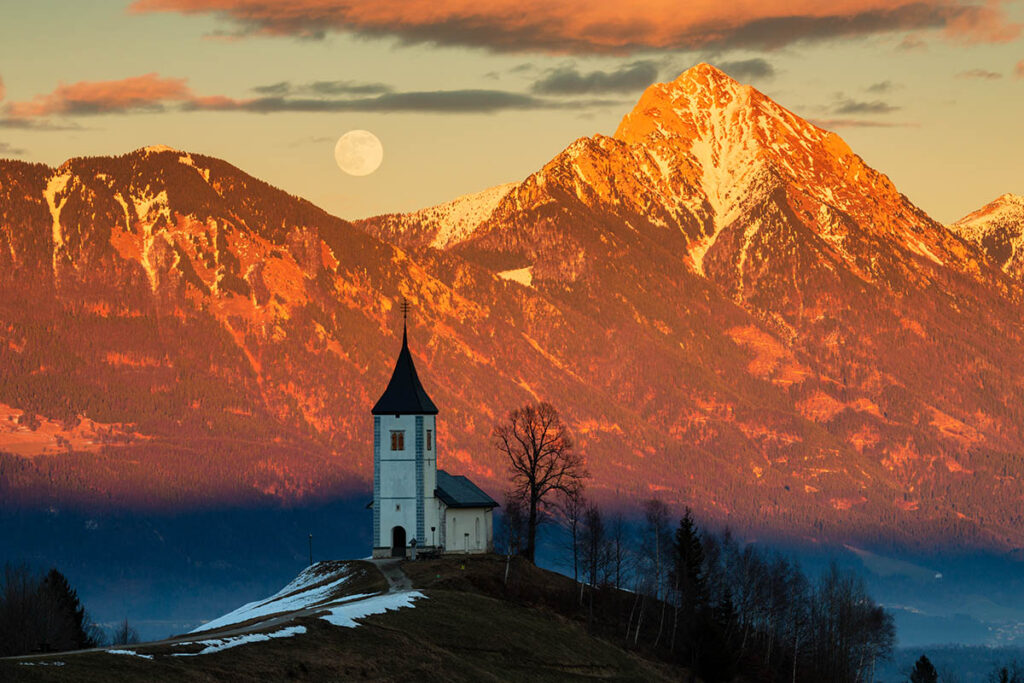 Full moon rising at sunset over Jamnik church of Saints Primus and Felician, perched on a hill on the Jelovica Plateau with the kamnik alps and Storzic mountain in the background, Slovenia.