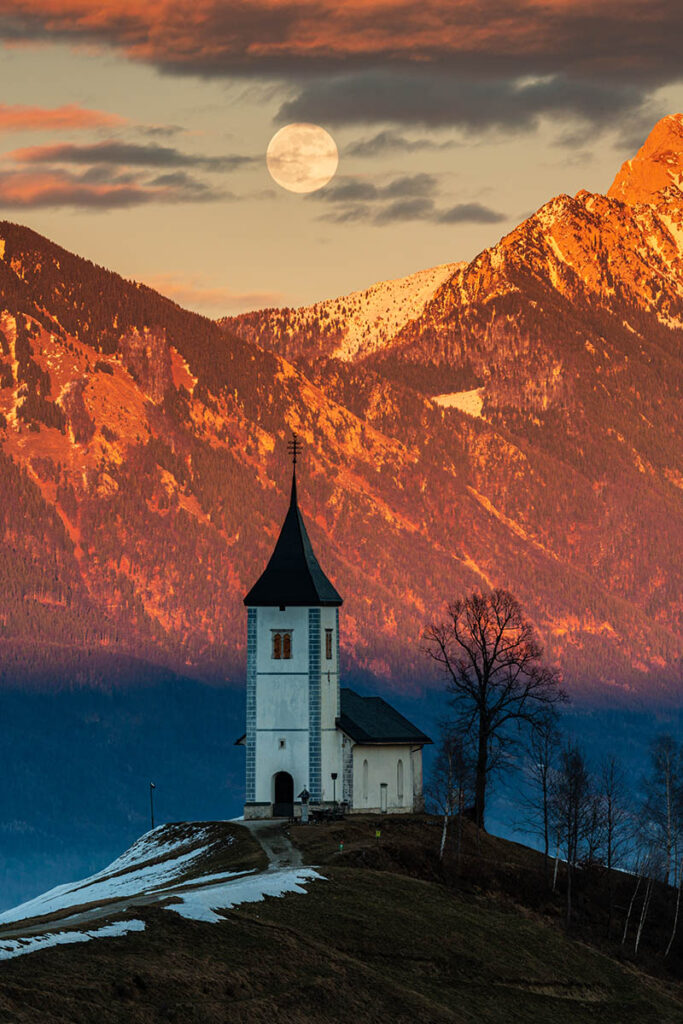 Full moon rising at sunset over Jamnik church of Saints Primus and Felician, perched on a hill on the Jelovica Plateau with the kamnik alps and Storzic mountain in the background, Slovenia.