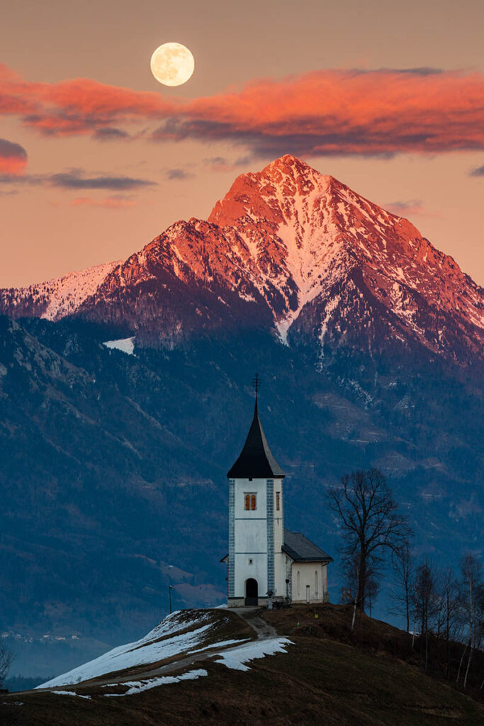 Full moon rising at sunset over Jamnik church of Saints Primus and Felician, perched on a hill on the Jelovica Plateau with the kamnik alps and Storzic mountain in the background, Slovenia.