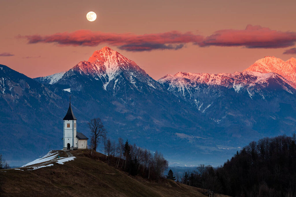 Full moon rising at sunset over Jamnik church of Saints Primus and Felician, perched on a hill on the Jelovica Plateau with the kamnik alps and Storzic mountain in the background, Slovenia.