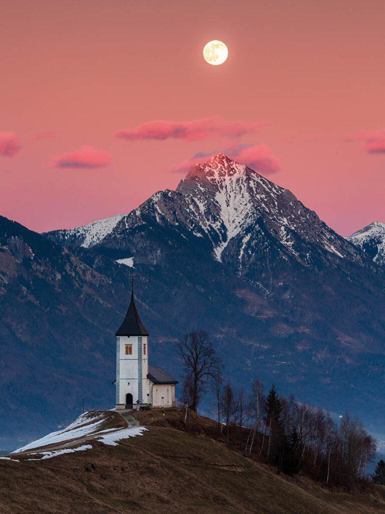 Full moon rising at sunset over Jamnik church of Saints Primus and Felician, perched on a hill on the Jelovica Plateau with the kamnik alps and Storzic mountain in the background, Slovenia.