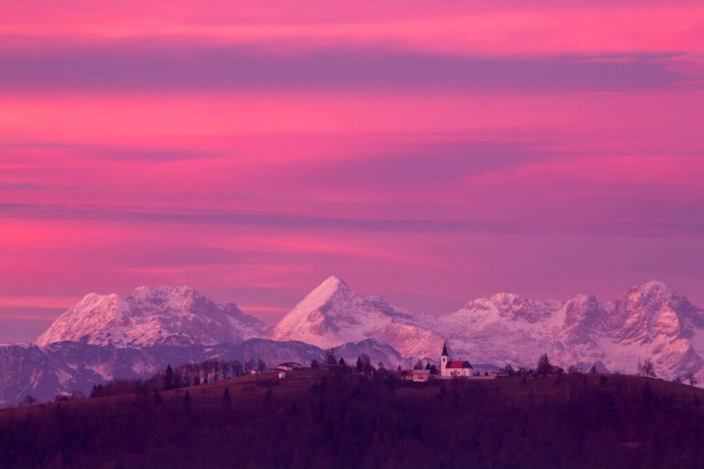 View across to the church of Saint Nicholas and the village of Jance at sunset with the Kamnik Alps in the background, in the hilly region to the east of Ljubljana, Slovenia. Seen from Mali Vrh village.