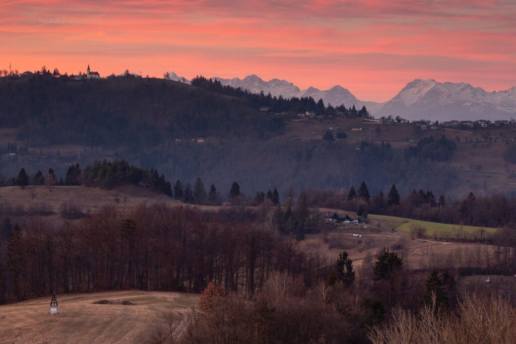 Sunset view across a hill in prezganje to the church of Saint Nicholas in the village of Jance and the Kamnik Alps to the east of Ljubljana, Slovenia. The shrine on the first hill is to Jesus Christ, built to commemorate the first visit of Pope John Paul the second to Slovenia in 1996.