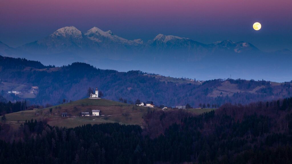 The full moon rising in winter over the mountains behind Sveti Tomaz nad Praprotnim (church of Saint Thomas) and the Kamnik Alps, Slovenia.