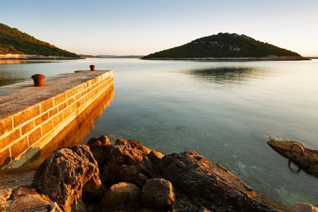 The morning sunrise casts warm light and colour over the rocky coastal area and pier of Cunski and casts a shadow over the island of Veli Osir. Cunski lies just 8kms north of Mali Losinj on Losinj Island, Croatia.