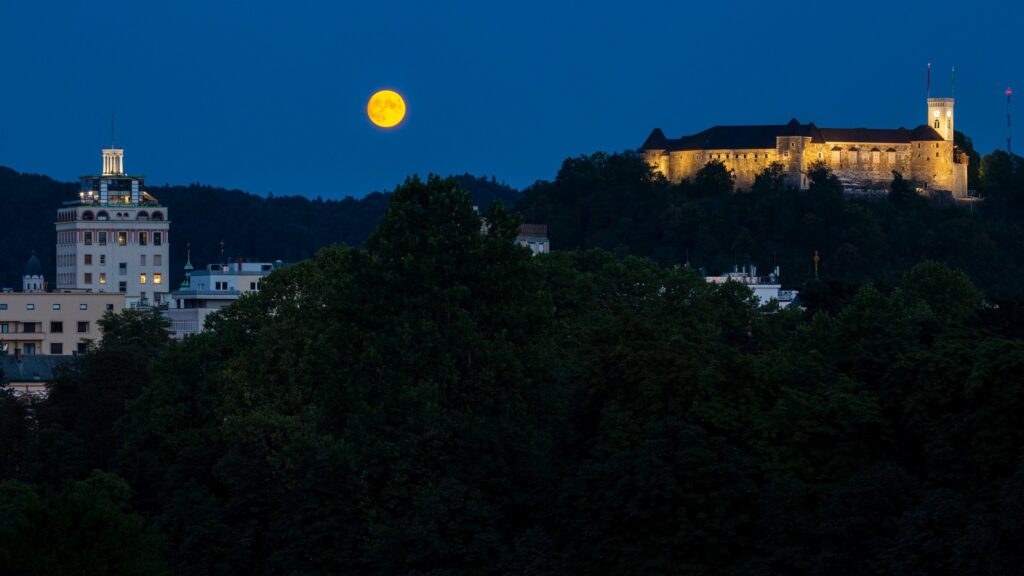 View across to the Ljubljana Castle as the full moon rises between it and Neboticnik Skyscraper at dusk. Seen from Tivoli Park, Ljubljana, Slovenia.