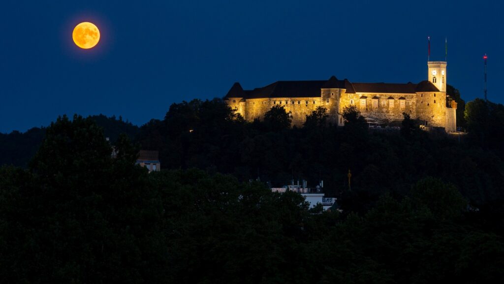 View across to the Ljubljana Castle as the full moon rises beside it at dusk. Seen from Tivoli Park, Ljubljana, Slovenia.