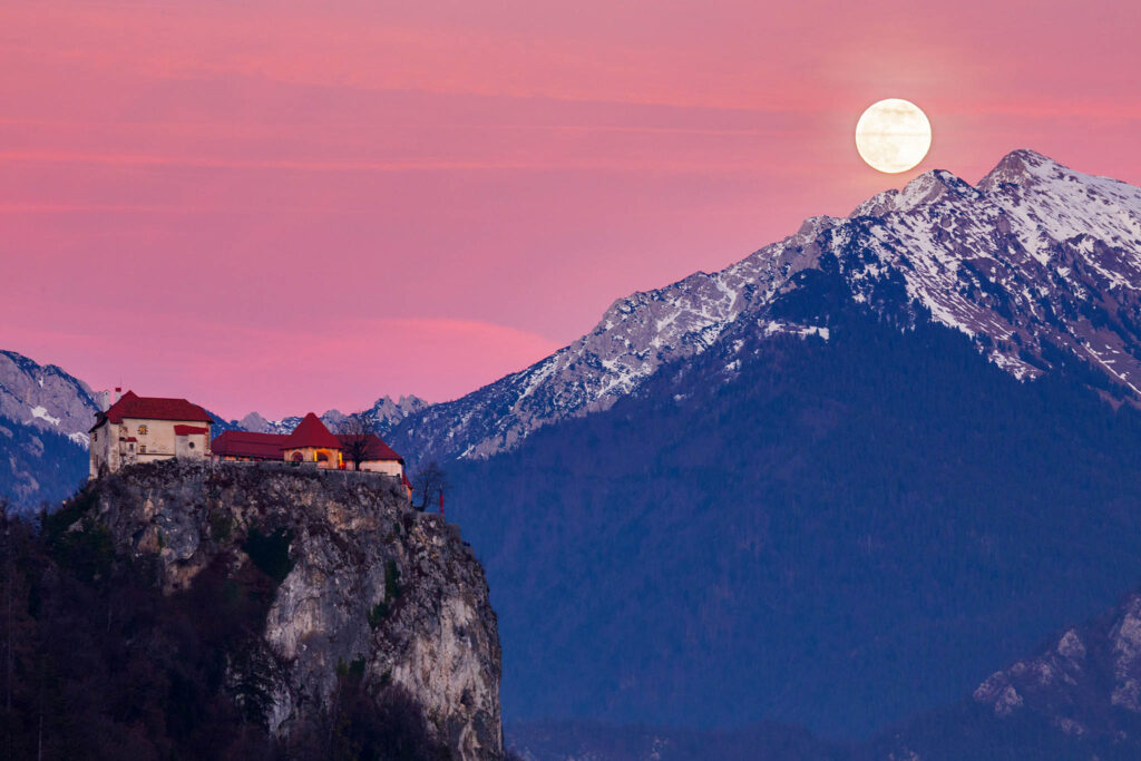 Full moon rising over Veliki Vrh mountain peak at sunset beside the castle at Lake Bled, Slovenia.