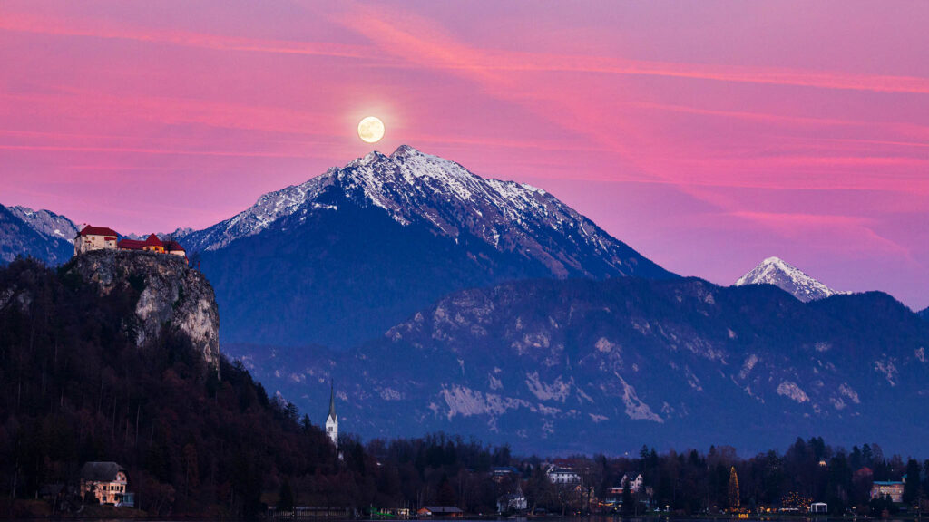 Full moon rising over Veliki Vrh mountain peak at sunset beside the castle at Lake Bled, Slovenia.