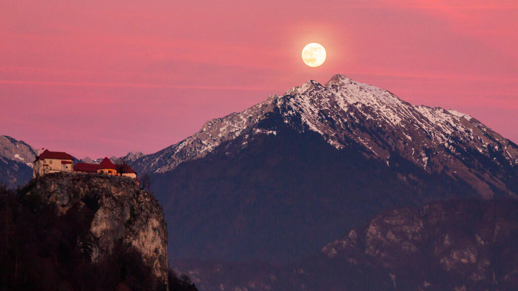 Full moon rising over Veliki Vrh mountain peak at sunset beside the castle at Lake Bled, Slovenia.