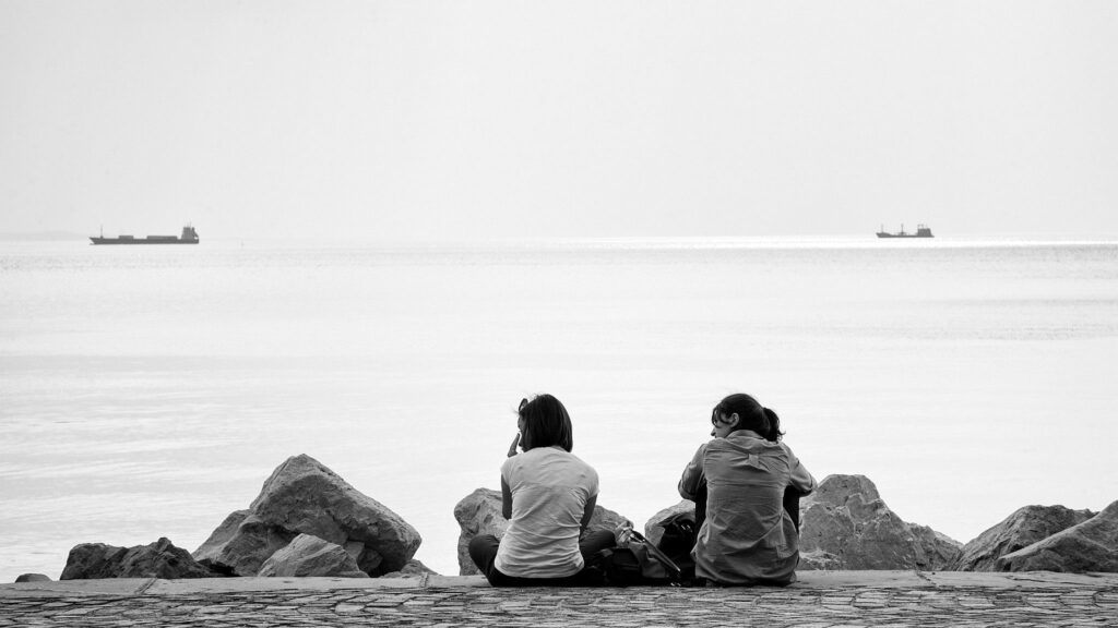 Two girls sitting on the promenade along viale miramare watching the ships go by in Trieste Bay, Italy.