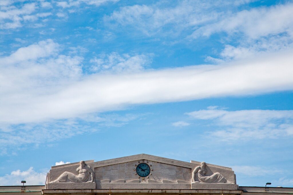 The clock and carvings on the Stazione Marittima building set against a lovely blue sky with wispy clouds, Trieste, Italy. The Stazione Marittima building is one of the greatest works carried out by the architects Umberto Nordio and Giacomo Zammattio in this wonderful little port town on the Adriatic. Aswell as being a passenger terminal for travellers boarding the many cruise ships that dock here, this building also houses the promotrieste convention and visitors bureau.