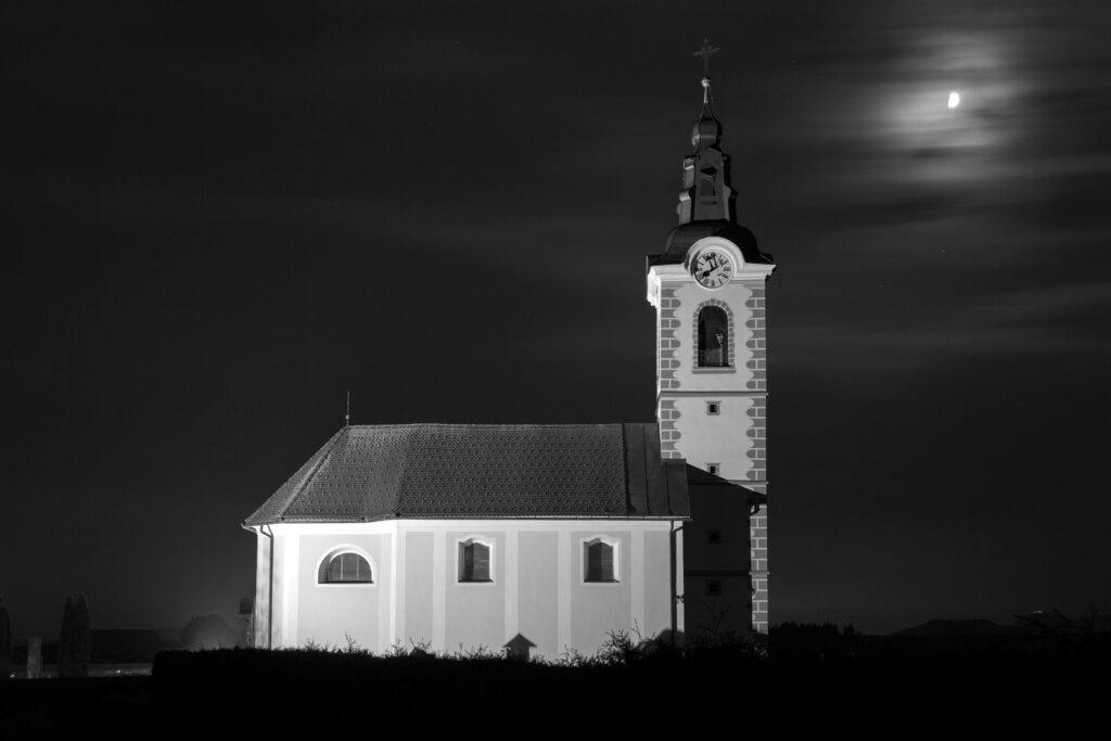 Black and white nightscape image of the church of saint John (Sveti Janez) just after dusk as clouds drift past the moon, Brnik, Slovenia. You can also see the church on Smarna Gora hill in the distance at the bottom right.