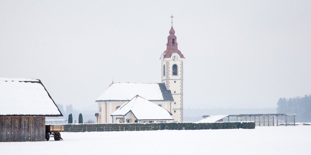 Winter view of the church of saint John as the snow falls, Brnik, near the Ljubljana airport, Slovenia. To the left is a farmer&#039;s hut and to the right is a Kozolec, a wooden rack for drying hay in the summer.