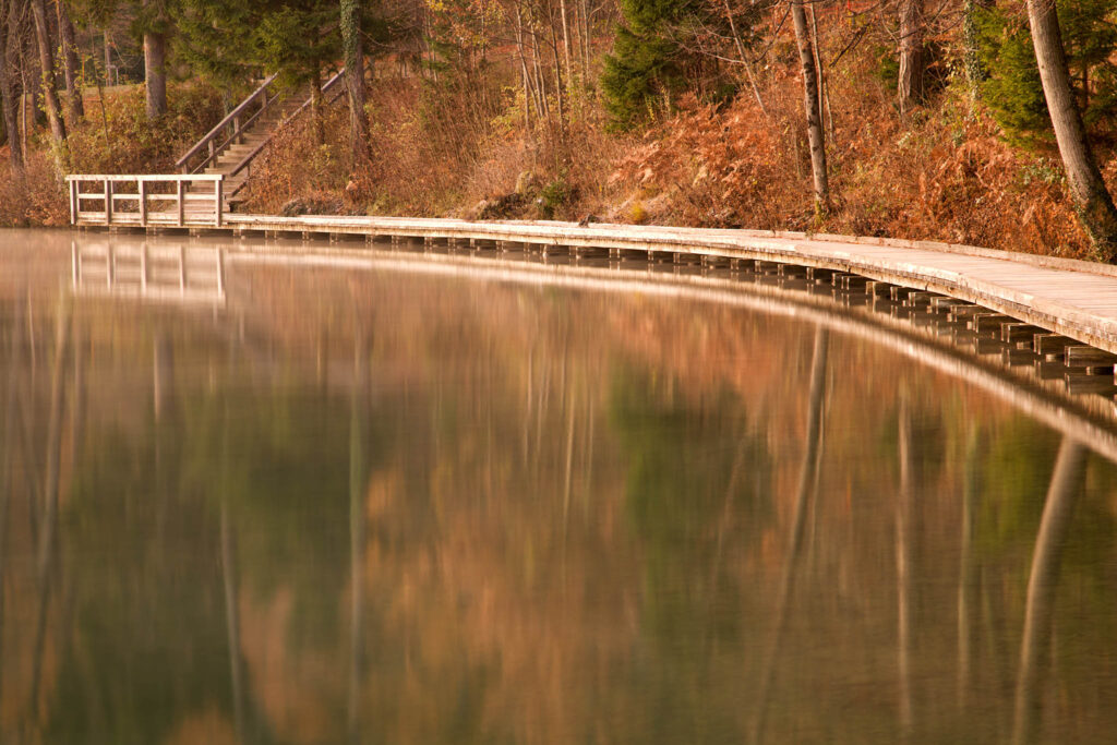 View along the wooden walkway beside Lake Bled with the autumn colours and trees reflected in the still water, Slovenia.