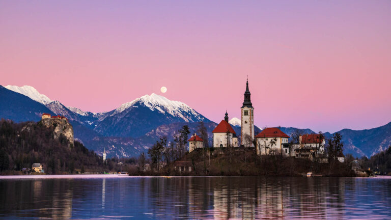 Full moon (January Wolf Moon) rising over Veliki Vrh mountain peak at sunset beside the island church at Lake Bled, Slovenia.