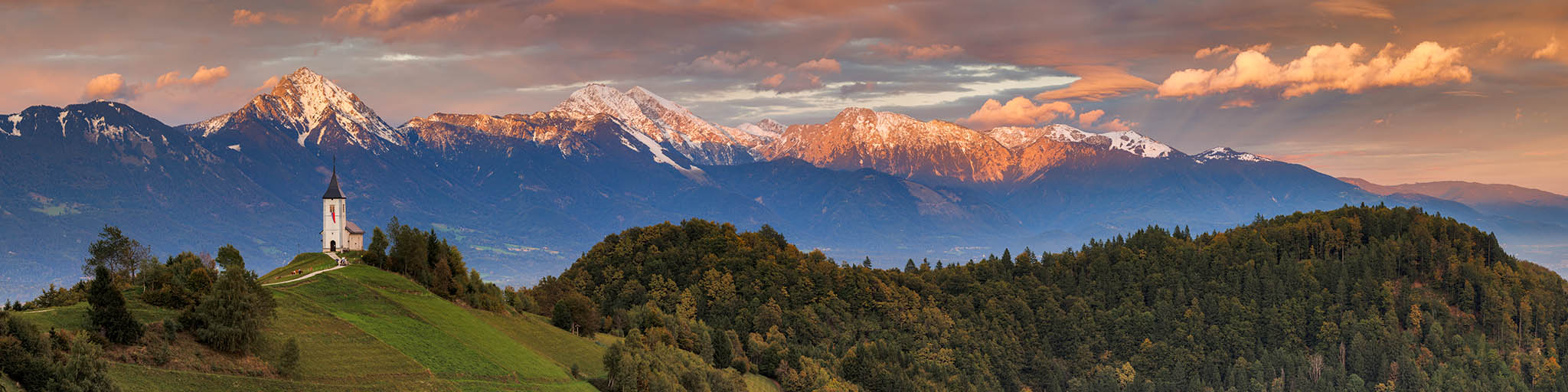 Stitched Panorama of sunset at Jamnik church of Saints Primus and Felician, perched on a hill on the Jelovica Plateau with the kamnik alps and Storzic mountain in the background, Slovenia.